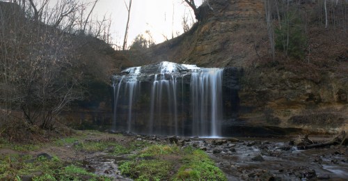 A serene waterfall cascades over rocky cliffs, surrounded by trees and lush greenery in a tranquil natural setting.