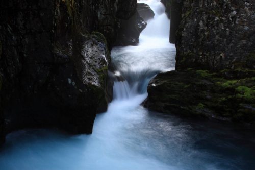 A serene waterfall flows between rocky cliffs, creating a misty, tranquil scene with smooth, blue water.