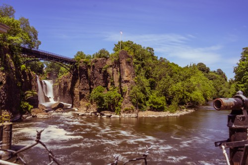 A scenic view of a waterfall surrounded by lush greenery, with a bridge overhead and a river flowing nearby.