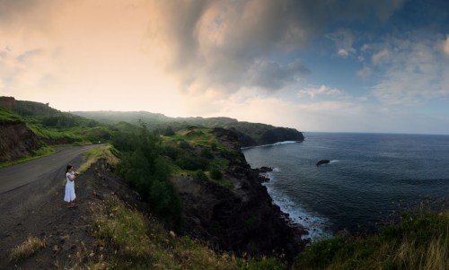 A person in a white dress stands on a cliff overlooking a serene ocean and lush green landscape under a cloudy sky.