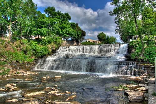 A serene waterfall cascades over rocks, surrounded by lush greenery and a partly cloudy sky.