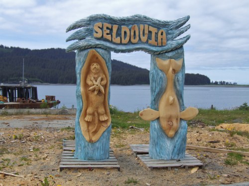 Wooden archway sign reading "Seldovia," featuring carved sea otter and fish designs, set against a coastal backdrop.