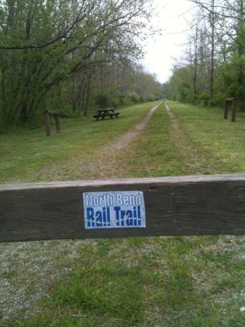 A grassy trail lined with trees, featuring a sign for the North Bend Rail Trail and picnic tables in the background.