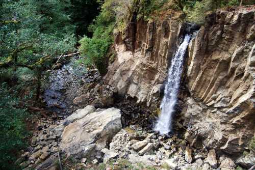 A waterfall cascading down rocky cliffs, surrounded by lush greenery and boulders in a serene natural setting.