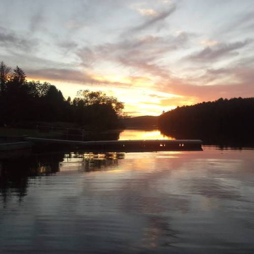 A serene lake at sunset, with a wooden dock extending into calm waters and trees silhouetted against the colorful sky.