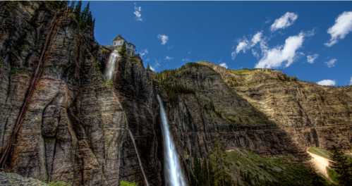 A scenic view of a rocky cliff with waterfalls cascading down, framed by lush greenery and a blue sky with clouds.