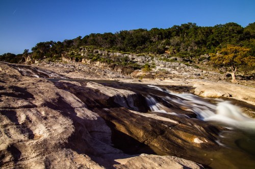 A serene river flows over smooth rocks, surrounded by lush greenery and a clear blue sky.