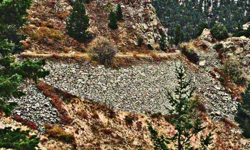 A rocky hillside with a winding path and sparse trees, showcasing a stone wall built into the landscape.