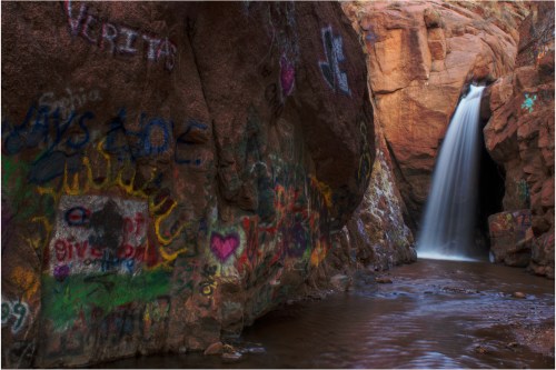 A waterfall cascades down rocky cliffs, surrounded by colorful graffiti on the rocks and a serene pool below.