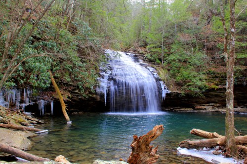 A serene waterfall cascades into a clear pool, surrounded by lush greenery and icicles hanging from rocks.