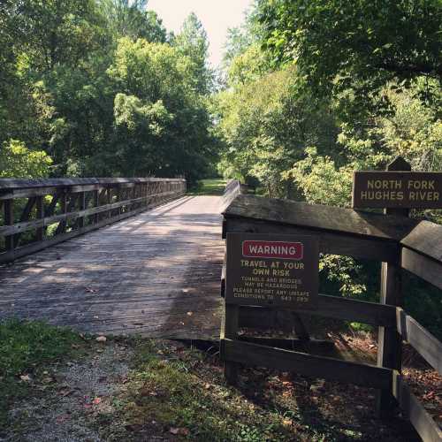 A wooden bridge leads to a path by the North Fork Hughes River, surrounded by lush greenery and warning signs.