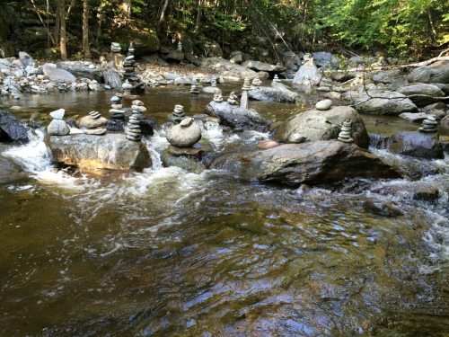 A serene river scene with stacked stones on rocks, surrounded by lush greenery and flowing water.