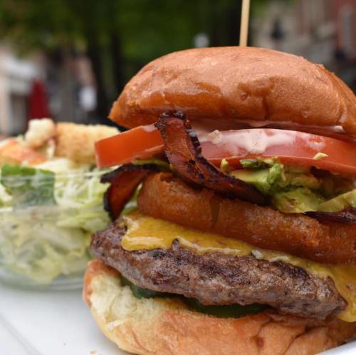 A close-up of a juicy burger with bacon, tomato, lettuce, and cheese, served with a side of salad.