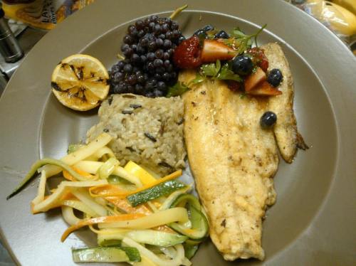 A plate featuring grilled fish, wild rice, sautéed vegetables, and fresh berries with lemon.