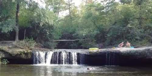 A serene scene of a waterfall with two people sitting on the rocks and one person swimming in the water.