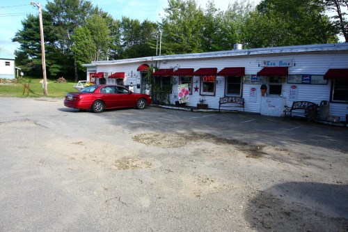 A red car parked in front of a small ice cream shop with a white exterior and red awnings, surrounded by trees.