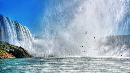 A stunning waterfall cascades into a misty pool, with birds flying through the spray under a clear blue sky.