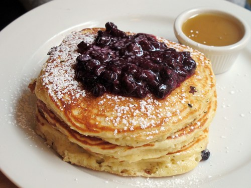 Stack of fluffy pancakes topped with berry compote and powdered sugar, served with a side of syrup.
