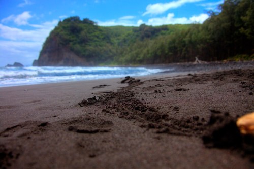 Footprints in dark sand lead to a lush green hillside and ocean waves under a blue sky with scattered clouds.