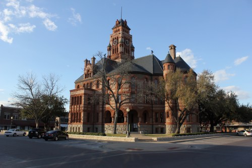 Historic red brick building with a tall clock tower, surrounded by trees and parked cars under a clear blue sky.
