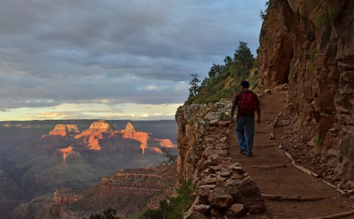 A hiker walks along a rocky trail at the Grand Canyon, with sunset light illuminating the distant cliffs.