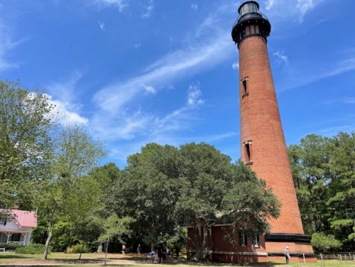 A tall brick lighthouse stands against a blue sky, surrounded by green trees and a small building nearby.