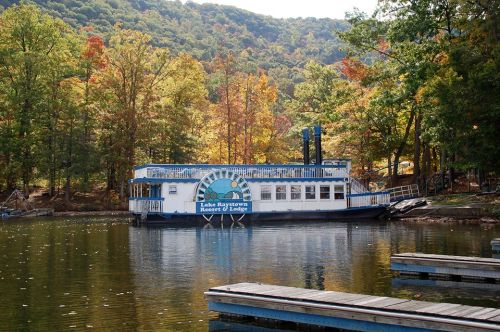A houseboat docked on a calm lake, surrounded by colorful autumn trees and mountains in the background.