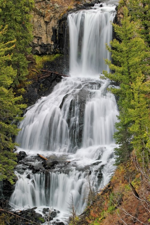 A cascading waterfall flows down rocky cliffs, surrounded by lush green trees and natural vegetation.