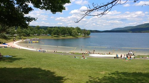 A scenic lake view with sandy shores, people swimming, and lush green trees under a partly cloudy sky.