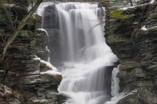 A serene waterfall cascading over rocky cliffs, surrounded by snow and greenery.