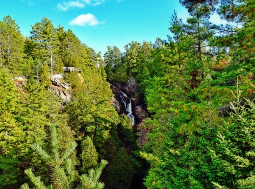 A scenic view of a waterfall surrounded by lush green trees under a blue sky with scattered clouds.