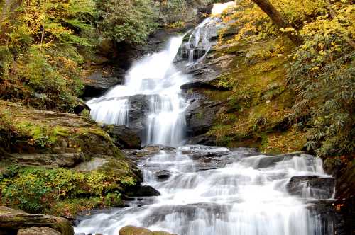 A serene waterfall cascades over rocky terrain, surrounded by lush greenery and autumn-colored foliage.