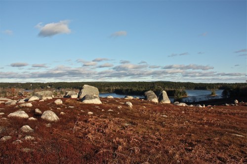 A scenic landscape featuring large boulders, a calm lake, and a clear blue sky with scattered clouds.