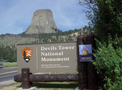 Sign for Devils Tower National Monument with the iconic tower in the background, surrounded by trees and a clear sky.