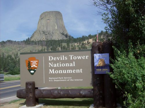 Sign for Devils Tower National Monument with the iconic tower in the background, surrounded by trees and a clear sky.