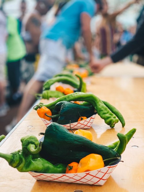 Colorful peppers and chilies arranged in baskets on a table, with people blurred in the background at an outdoor event.
