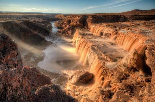 A stunning waterfall cascades over rocky cliffs into a river, illuminated by sunlight in a vast, arid landscape.