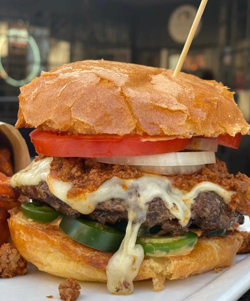Close-up of a juicy burger with melted cheese, jalapeños, tomato, and onion on a toasted bun, served with fries.