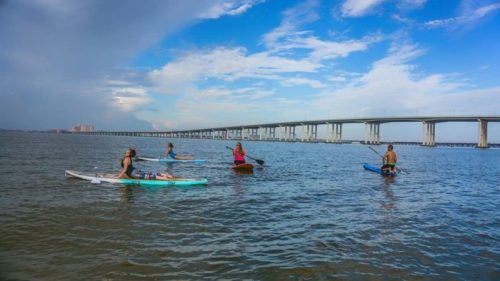 Four people paddleboarding on calm water under a blue sky with clouds, near a long bridge in the background.