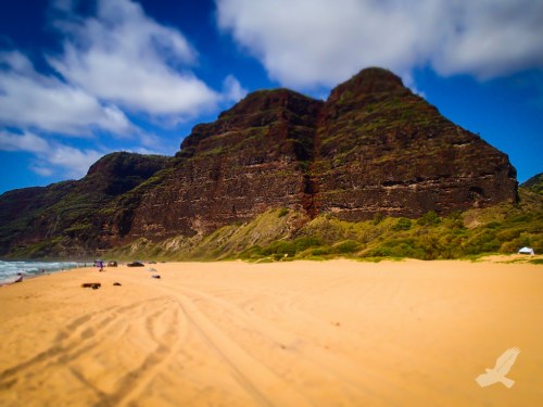 A sandy beach with a rocky, green-covered mountain in the background under a blue sky with clouds.