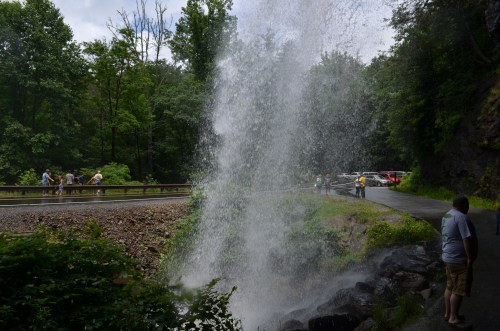 A waterfall cascades down rocks near a road, with people observing and lush greenery surrounding the scene.