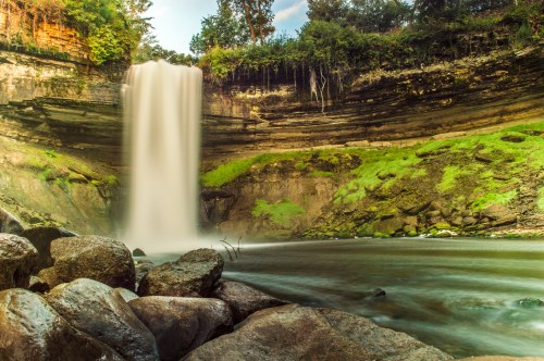 A serene waterfall cascades over rocky cliffs into a calm river, surrounded by lush greenery and smooth stones.