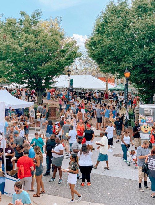 A bustling outdoor festival with large crowds, food stalls, and tents under green trees on a sunny day.