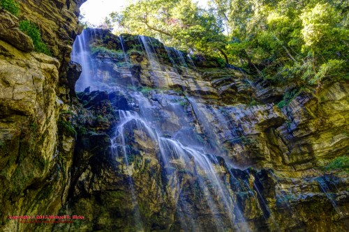A cascading waterfall flows down a rocky cliff surrounded by lush greenery and sunlight filtering through the trees.