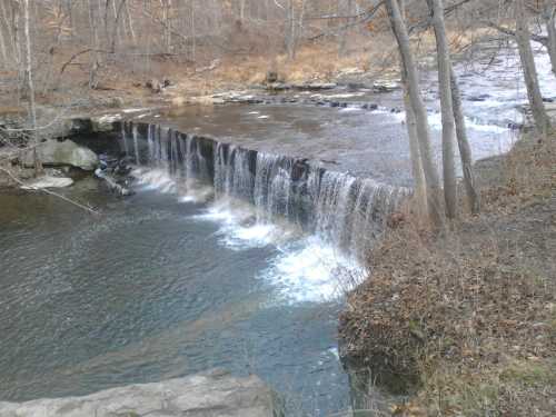 A serene waterfall cascading over rocks into a calm river, surrounded by bare trees and autumn foliage.