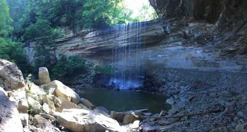 A serene waterfall cascades over rocky cliffs into a calm pool, surrounded by lush greenery and rugged terrain.