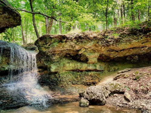 A serene waterfall cascades over rocky terrain, surrounded by lush green trees in a peaceful forest setting.