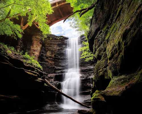 A serene waterfall cascades down rocky cliffs, surrounded by lush greenery and a bridge overhead.