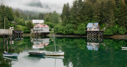 A serene waterfront scene with colorful houses on stilts, surrounded by lush trees and calm water reflecting the landscape.