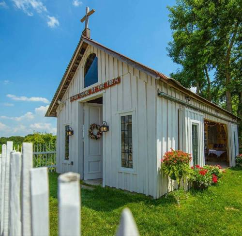 A charming white chapel with a cross on top, surrounded by greenery and flowers, under a clear blue sky.